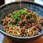 A bowl of Sticky Beef Noodles with tender beef, glossy sauce, and fresh garnishes.